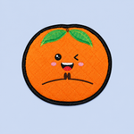An embroidered round orange patch with a winking face, green leaves, pink cheeks, and a black stitched border on a light blue background.